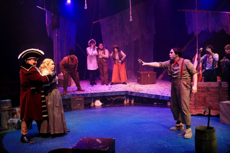 Scene in a play of a pirate holding a young lady hostage while a young man reaches out to her.